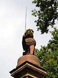 Ein marmorner Lwe auf einer hohen Sule, in der rechten Klaue ein bloes Schwert, auf dem Kopf eine Sturmhaube tragend.